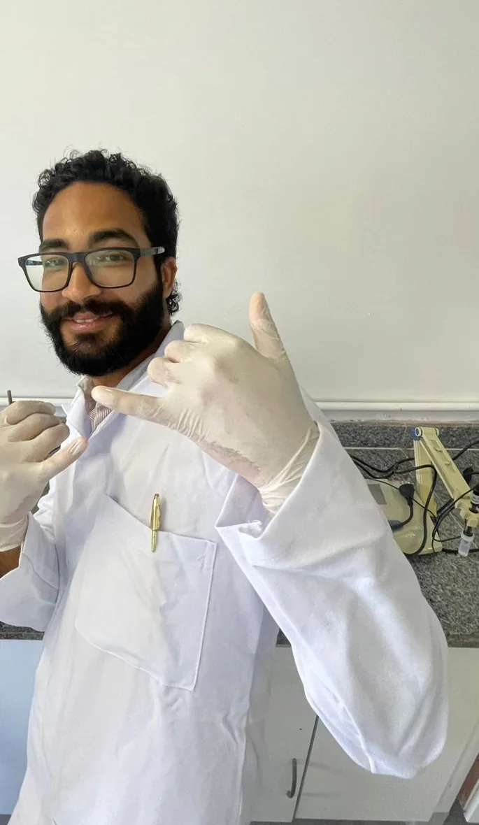 Daniel P Francisco sorrindo, de jaleco e luvas, na bancada do laboratório