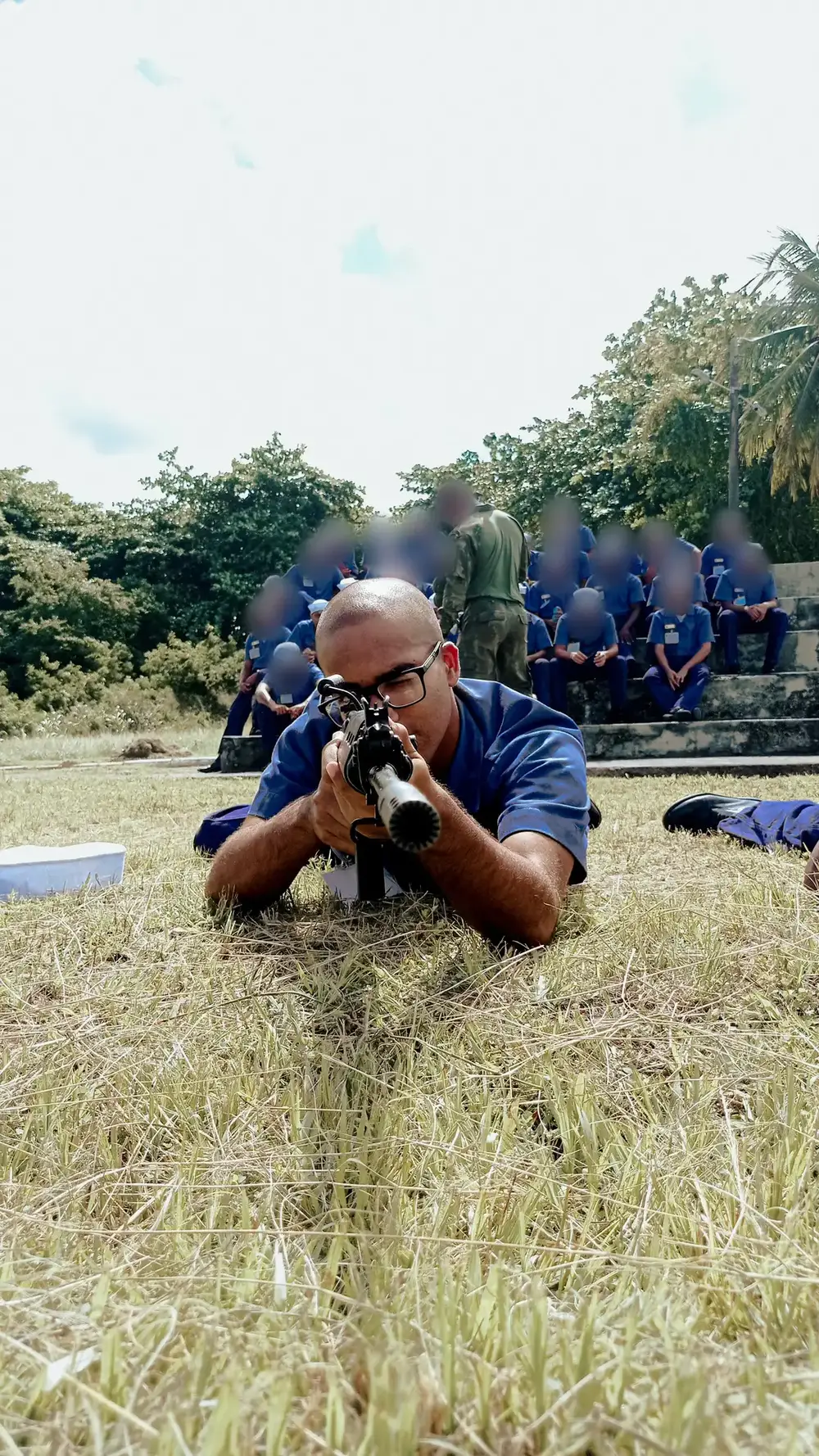 Daniel P Francisco lying on the grass in a firing position during military training, with the faces of people in the background blurred